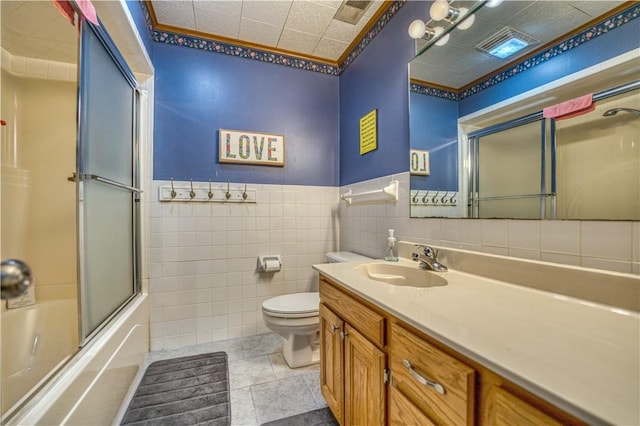 full bathroom featuring a wainscoted wall, tile walls, bath / shower combo with glass door, toilet, and vanity