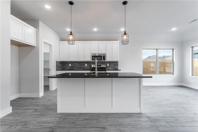 kitchen featuring white cabinetry, dark stone counters, a center island with sink, pendant lighting, and appliances with stainless steel finishes