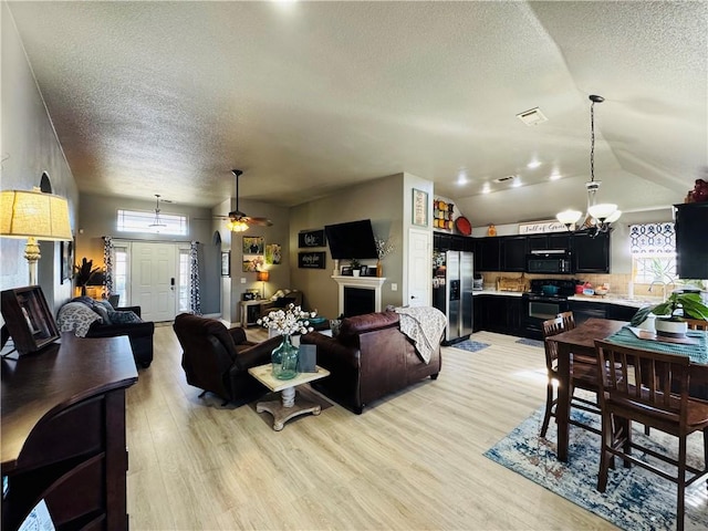 living area with visible vents, light wood-type flooring, vaulted ceiling, ceiling fan with notable chandelier, and a textured ceiling