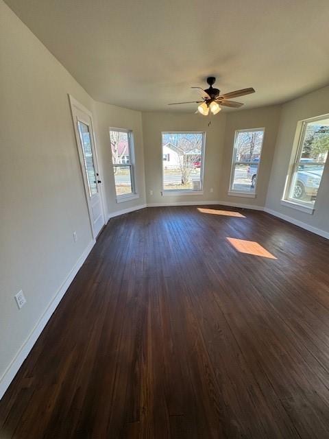 empty room with a healthy amount of sunlight, a ceiling fan, dark wood-type flooring, and baseboards