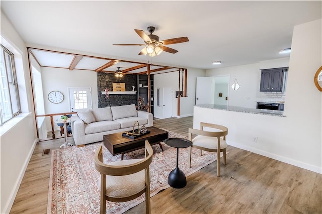 living room featuring ceiling fan, light hardwood / wood-style flooring, a stone fireplace, and lofted ceiling with beams