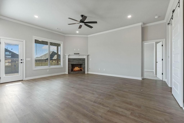 unfurnished living room featuring ceiling fan, a barn door, crown molding, a stone fireplace, and dark hardwood / wood-style flooring