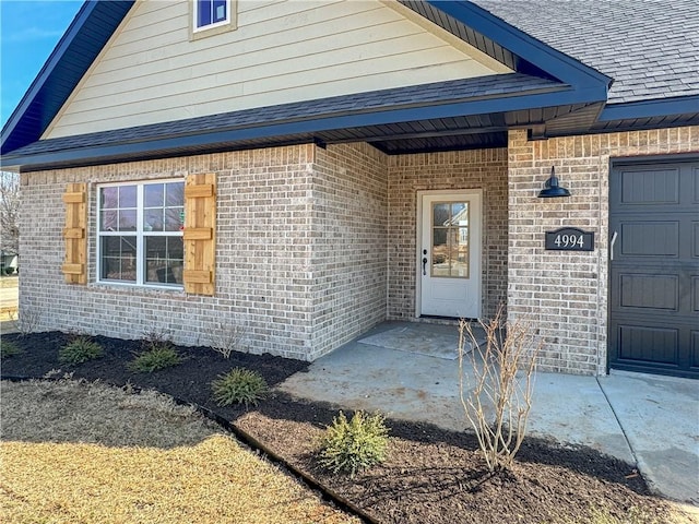 entrance to property with brick siding, a garage, and roof with shingles