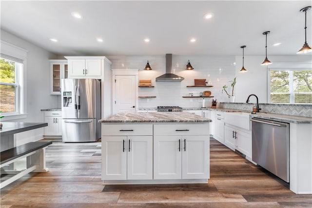 kitchen with white cabinets, backsplash, hanging light fixtures, stainless steel appliances, and wall chimney range hood