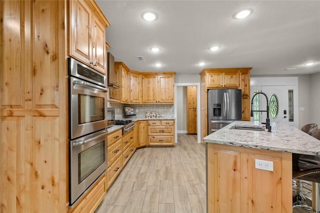 kitchen with appliances with stainless steel finishes, a breakfast bar area, a kitchen island with sink, light brown cabinets, and light hardwood / wood-style flooring