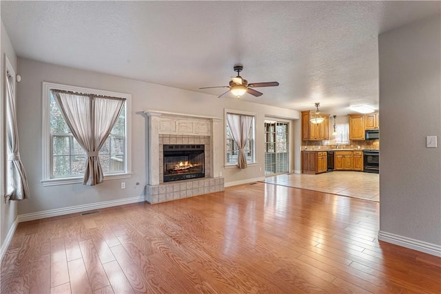 unfurnished living room featuring a ceiling fan, light wood finished floors, baseboards, a textured ceiling, and a tiled fireplace