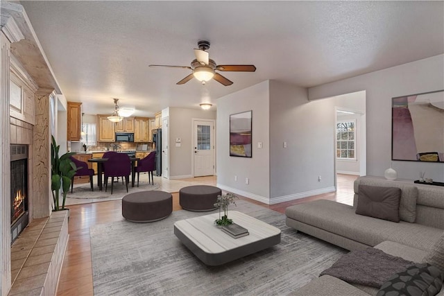 living room with a warm lit fireplace, a ceiling fan, light wood-style floors, baseboards, and a textured ceiling