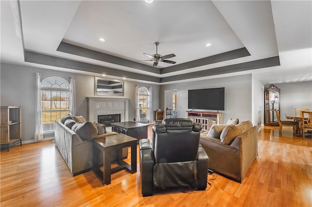 living room featuring ceiling fan, a fireplace, baseboards, light wood-style floors, and a tray ceiling