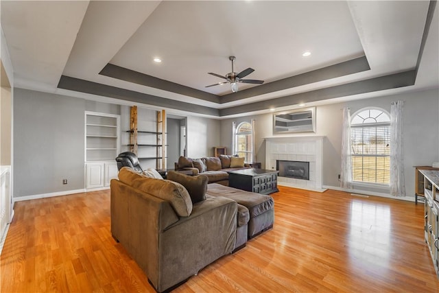 living room featuring light wood-style floors, a tray ceiling, baseboards, and a tile fireplace