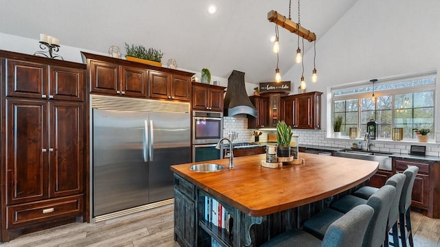 kitchen with decorative light fixtures, sink, custom exhaust hood, a kitchen island with sink, and stainless steel appliances