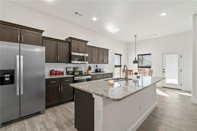 kitchen featuring an island with sink, sink, hanging light fixtures, light stone counters, and stainless steel appliances