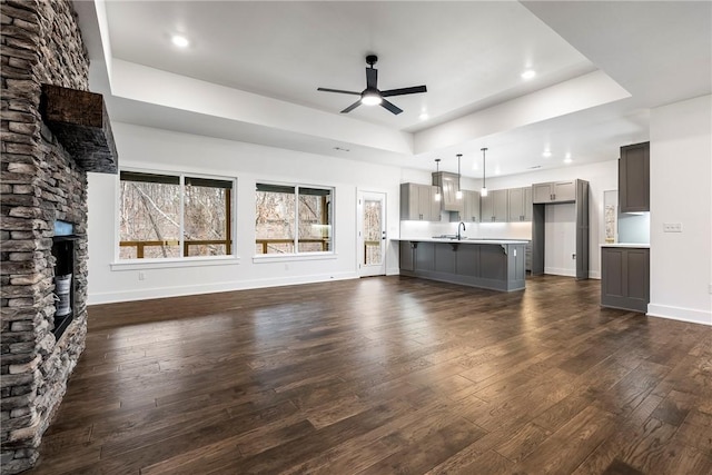 unfurnished living room with sink, dark wood-type flooring, ceiling fan, a stone fireplace, and a raised ceiling