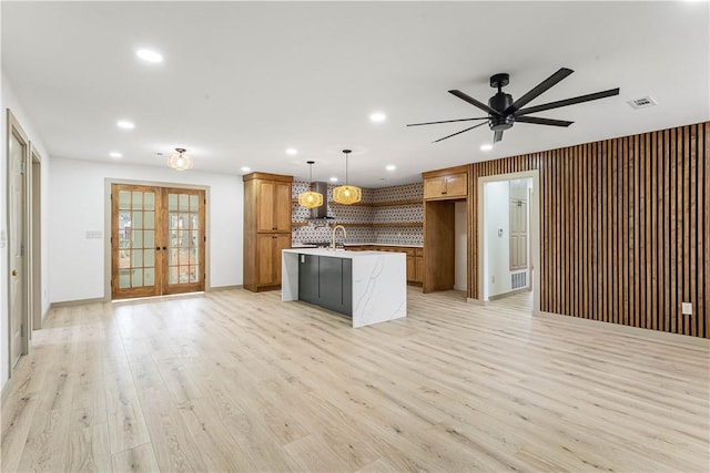 kitchen featuring french doors, tasteful backsplash, light wood-type flooring, pendant lighting, and a kitchen island with sink