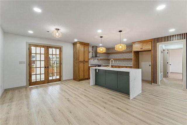 kitchen with pendant lighting, wall chimney range hood, tasteful backsplash, an island with sink, and french doors