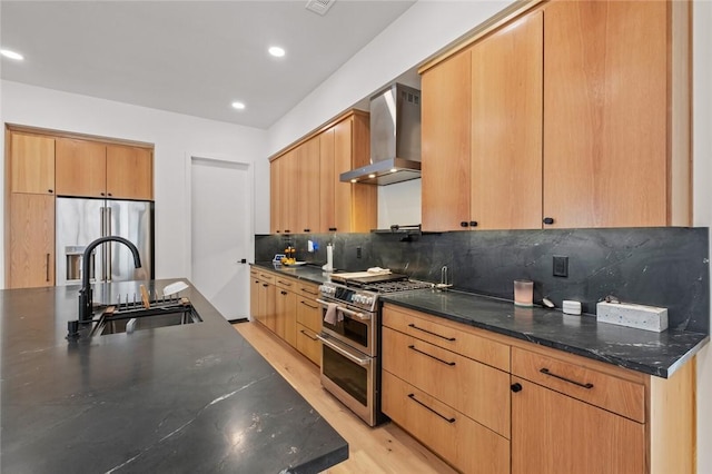 kitchen featuring sink, wall chimney range hood, dark stone countertops, stainless steel appliances, and backsplash