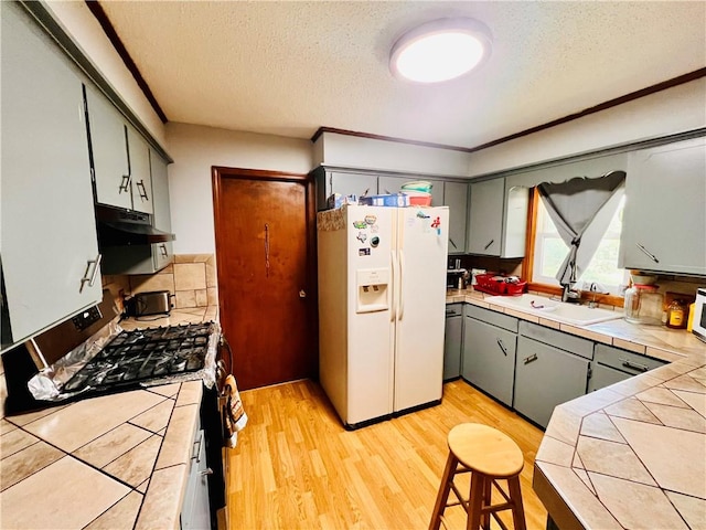 kitchen featuring a textured ceiling, light wood-type flooring, stainless steel range with gas cooktop, tile counters, and white fridge with ice dispenser