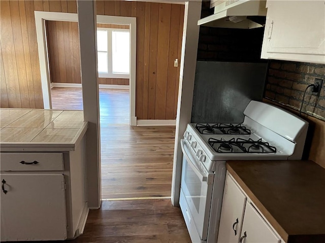 kitchen with dark wood-type flooring, white gas stove, white cabinets, and wood walls