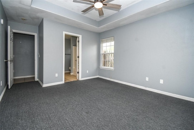 unfurnished bedroom with a raised ceiling, dark colored carpet, ensuite bathroom, baseboards, and ceiling fan