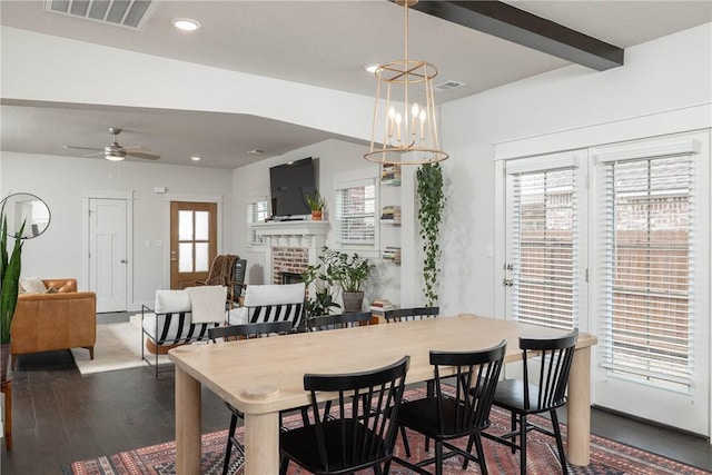 dining area with dark wood-type flooring, a fireplace, ceiling fan with notable chandelier, and beamed ceiling