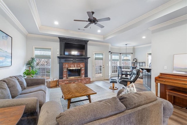 living room with light wood-type flooring, ornamental molding, a tray ceiling, ceiling fan, and a fireplace