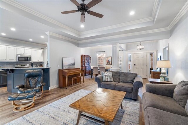 living room with ornate columns, ceiling fan with notable chandelier, a raised ceiling, crown molding, and light hardwood / wood-style flooring