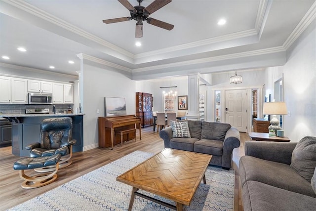 living room featuring decorative columns, ornamental molding, a tray ceiling, light wood-style floors, and ceiling fan with notable chandelier