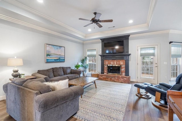 living area with ceiling fan, ornamental molding, hardwood / wood-style floors, a tray ceiling, and a brick fireplace