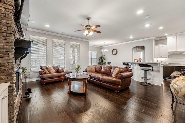 living room with ornamental molding, dark hardwood / wood-style flooring, ceiling fan with notable chandelier, and a stone fireplace