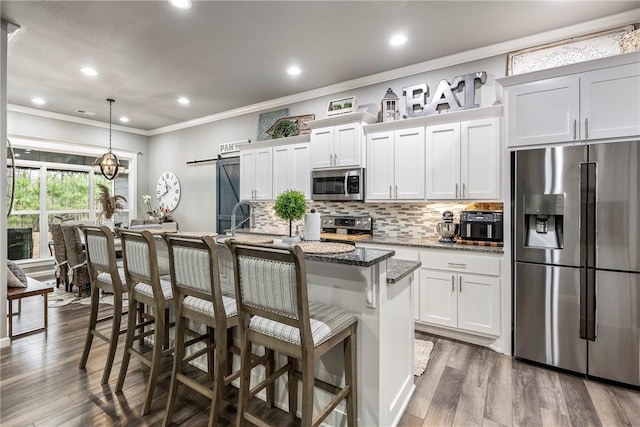 kitchen featuring a center island with sink, dark stone counters, stainless steel appliances, a barn door, and white cabinets