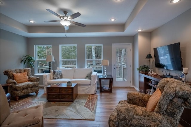 living area featuring a raised ceiling, wood finished floors, baseboards, and ceiling fan