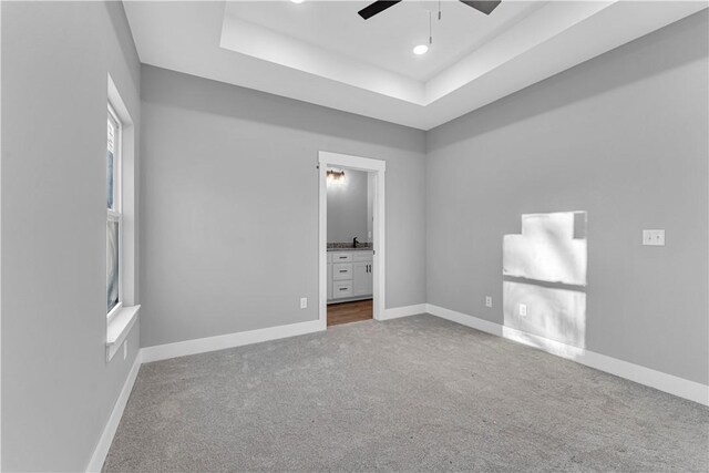 unfurnished bedroom with a raised ceiling, ensuite bath, carpet, baseboards, and ceiling fan
