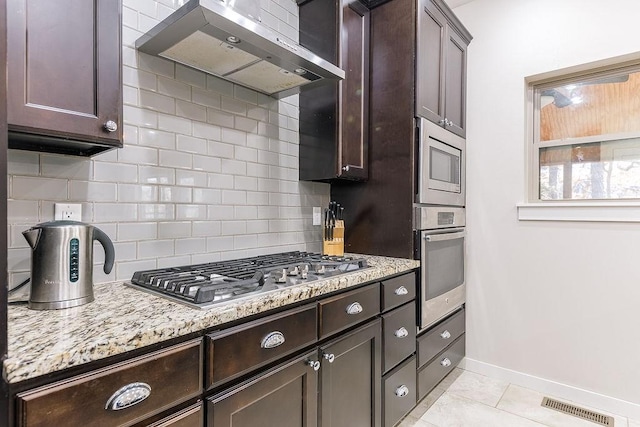 kitchen featuring appliances with stainless steel finishes, backsplash, light stone counters, dark brown cabinets, and wall chimney exhaust hood