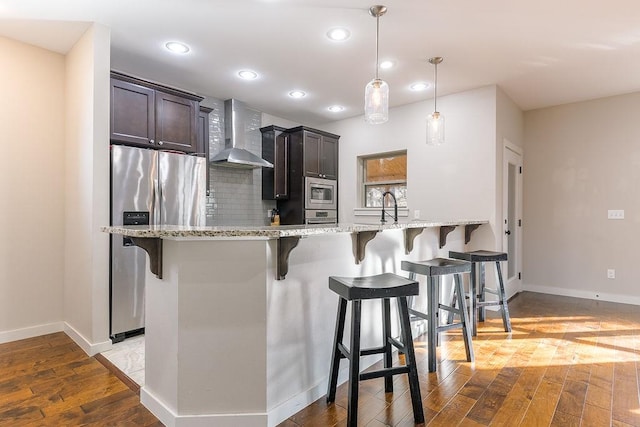 kitchen featuring appliances with stainless steel finishes, a kitchen breakfast bar, decorative light fixtures, wall chimney exhaust hood, and light wood-type flooring