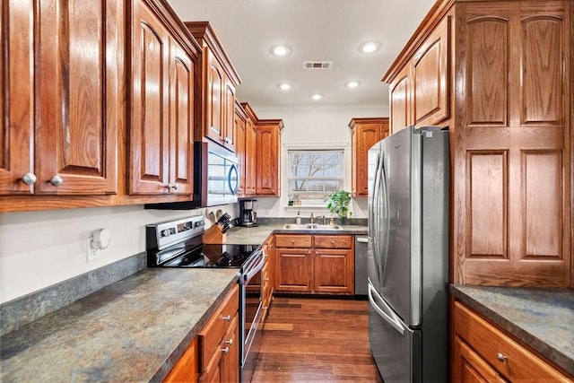 kitchen with appliances with stainless steel finishes, a sink, brown cabinetry, dark countertops, and dark wood-style floors