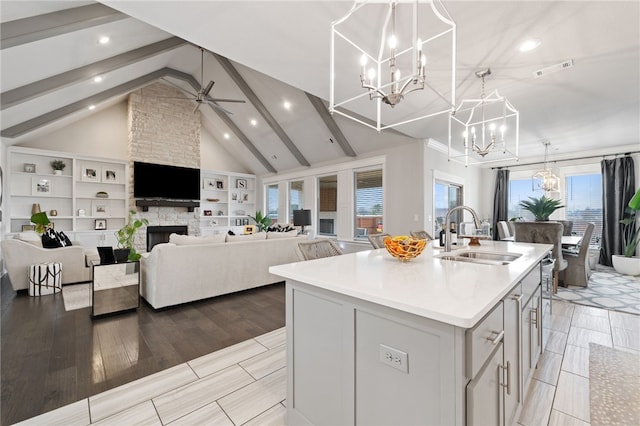 kitchen with a stone fireplace, beamed ceiling, sink, hanging light fixtures, and a kitchen island with sink