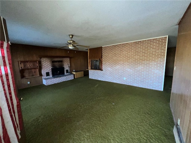 unfurnished living room with a brick fireplace, dark colored carpet, wood walls, and ceiling fan