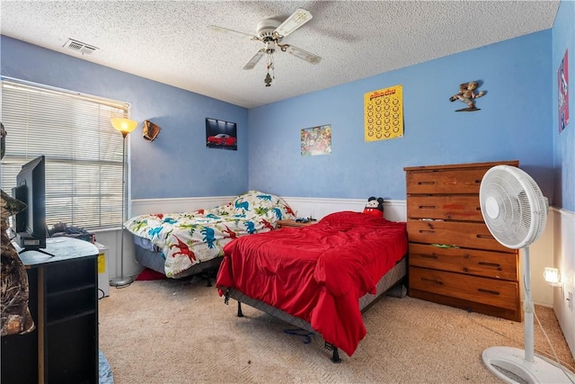 bedroom featuring light carpet, visible vents, a ceiling fan, a wainscoted wall, and a textured ceiling