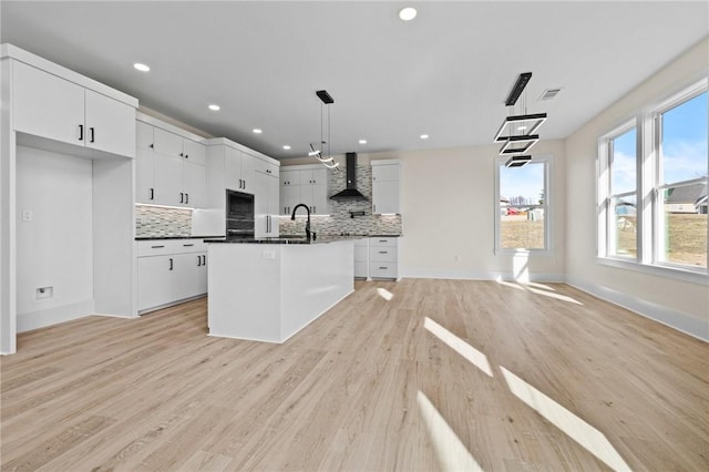 kitchen with dark countertops, hanging light fixtures, white cabinets, a kitchen island with sink, and wall chimney range hood