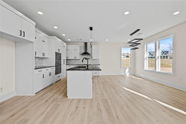 kitchen featuring white cabinets, an island with sink, hanging light fixtures, built in microwave, and wall chimney range hood