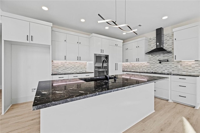 kitchen featuring stainless steel microwave, hanging light fixtures, a kitchen island with sink, white cabinets, and wall chimney range hood