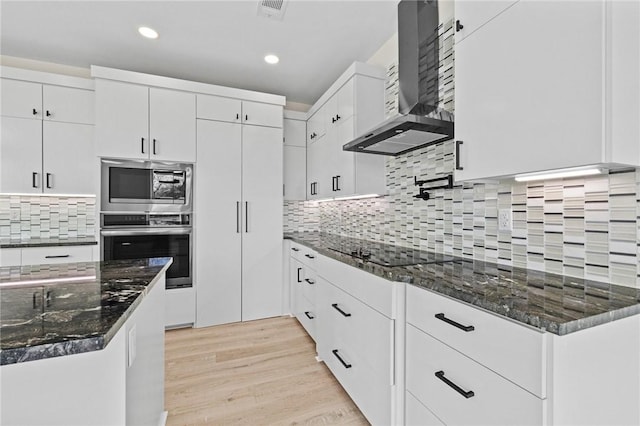 kitchen featuring light wood finished floors, appliances with stainless steel finishes, white cabinets, wall chimney range hood, and dark stone counters