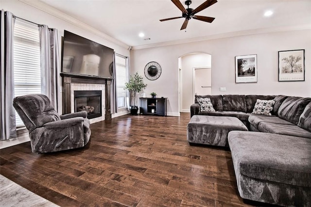 living room with ceiling fan, arched walkways, dark wood-type flooring, ornamental molding, and a tiled fireplace