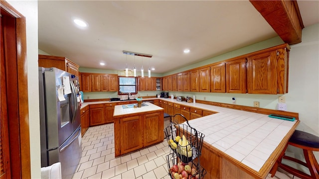 kitchen with brown cabinets, stainless steel refrigerator with ice dispenser, tile counters, recessed lighting, and a kitchen island