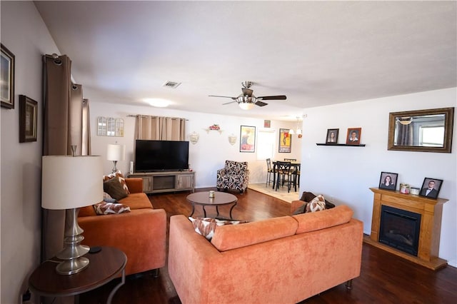 living area with dark wood-style floors, a glass covered fireplace, visible vents, and ceiling fan