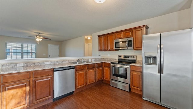 kitchen with stainless steel appliances, brown cabinetry, dark wood-type flooring, a sink, and light stone countertops