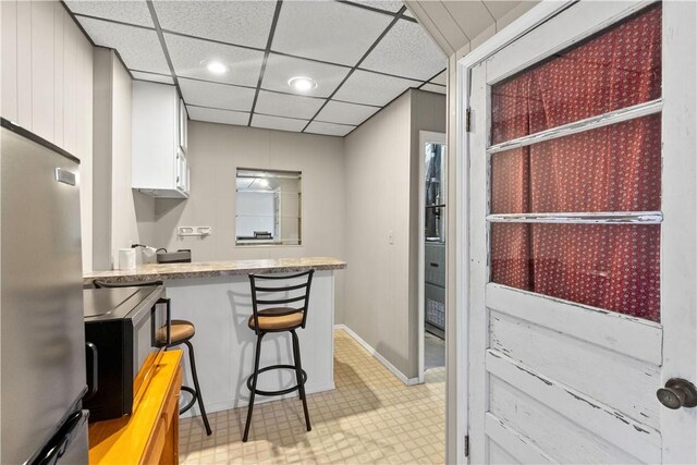 kitchen with light floors, a drop ceiling, freestanding refrigerator, white cabinets, and a kitchen bar