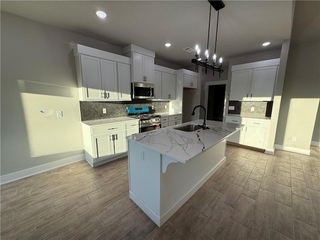 kitchen featuring a kitchen island with sink, a sink, white cabinets, appliances with stainless steel finishes, and pendant lighting