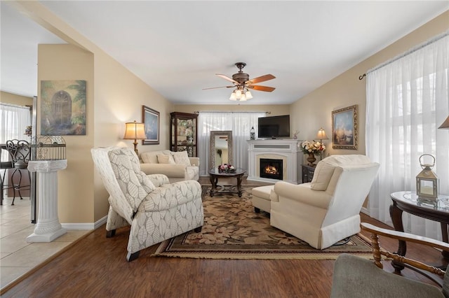 living room featuring a warm lit fireplace, ceiling fan, wood finished floors, and baseboards