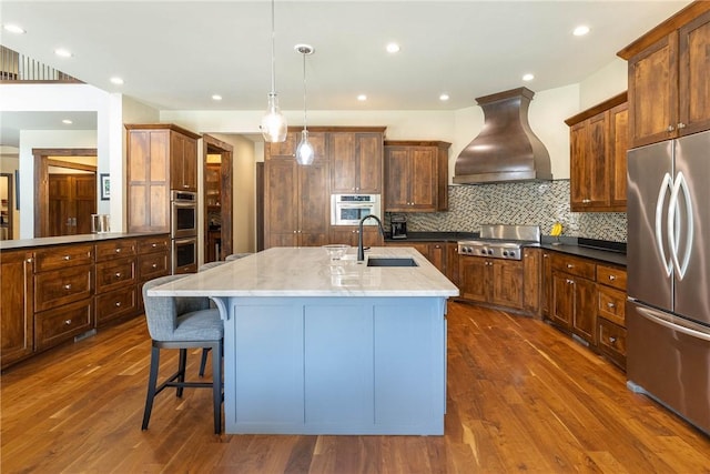 kitchen featuring a sink, hanging light fixtures, appliances with stainless steel finishes, custom exhaust hood, and a center island with sink