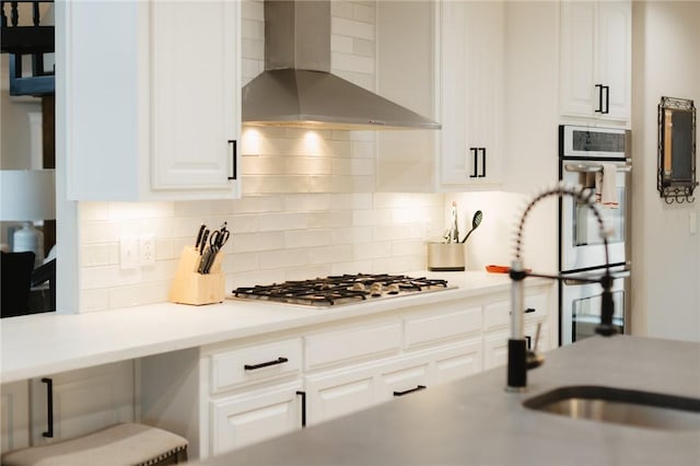 kitchen featuring decorative backsplash, wall chimney exhaust hood, white cabinetry, a sink, and stainless steel appliances